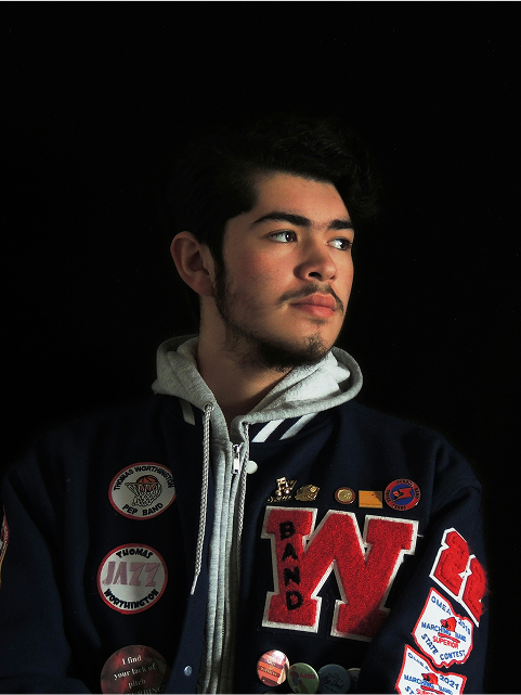 A young man wearing a letter-jacket with patches, looking off to the side against a dark background.